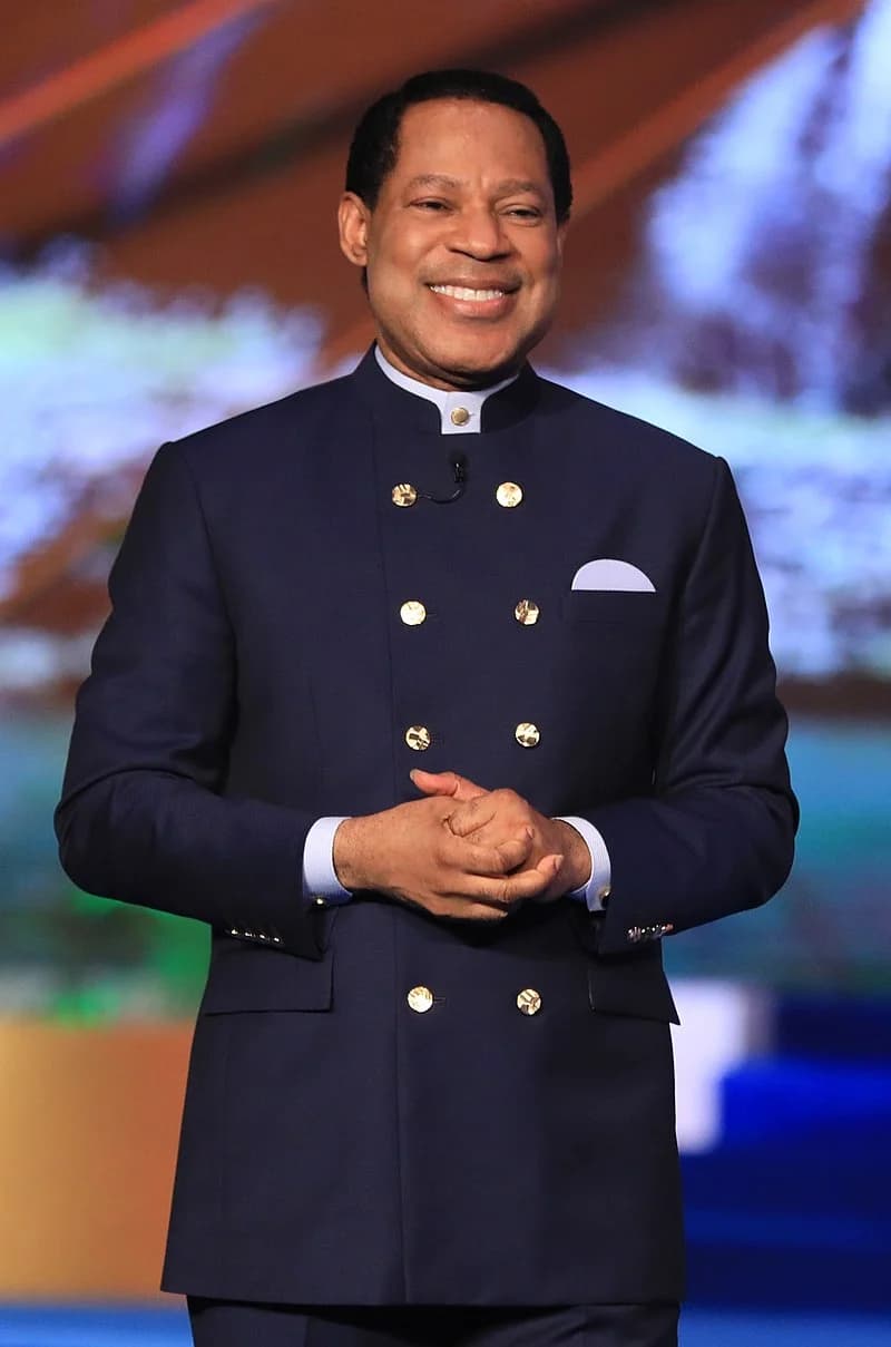 Pastor Chris Oyakhilome on stage, smiling, in a navy double-breasted jacket